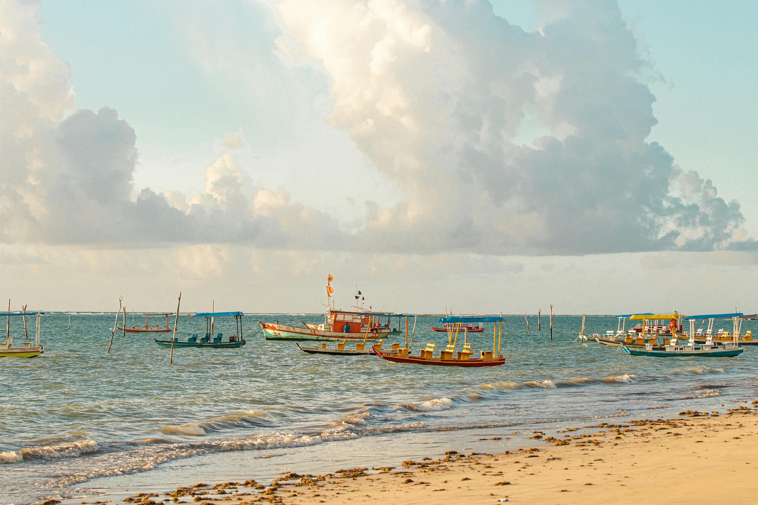 Vibrant fishing boats anchored near the sandy shore of São Miguel dos Milagres, Brazil under a partly cloudy sky.